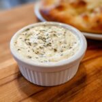 Close-up of creamy white pizza sauce seasoned with herbs in a small white ramekin, with pizza blurred in the background.