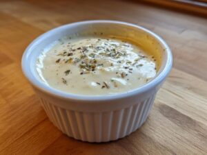A small white ramekin filled with creamy white pizza sauce, topped with dried herbs and spices.