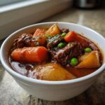 A close-up of a white bowl filled with rich, dark crockpot beef stew featuring tender beef chunks, large carrots, potatoes, and bright green peas.