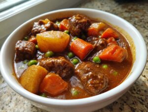A close-up of a white bowl filled with rich, dark crockpot beef stew featuring tender beef chunks, potatoes, carrots, and peas.