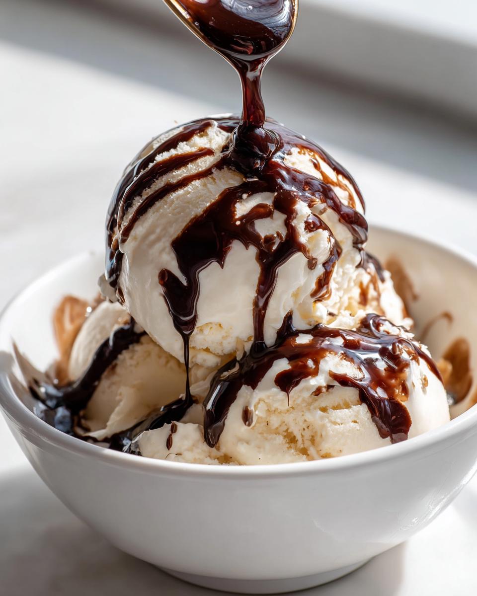 A spoonful of rich chocolate syrup being drizzled over scoops of vanilla ice cream in a white bowl.