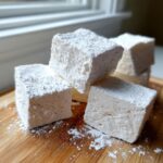 Close-up of several fluffy homemade marshmallows stacked on a wooden board, heavily dusted with powdered sugar.