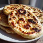 A stack of freshly cooked, slightly charred naan bread resting on a white plate.