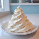 A tall, perfectly piped swirl of fluffy vanilla buttercream frosting sitting on a small white plate.