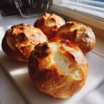 Four freshly baked, golden brown bread bowls with cracked tops resting on a white cutting board near a window.