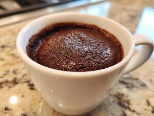 Close-up of a freshly baked, dark chocolate fudgy mug cake rising slightly above the rim of a white ceramic cup.