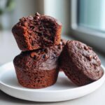 A stack of three rich, dark chocolate zucchini muffins topped with melted chocolate chips on a white plate.