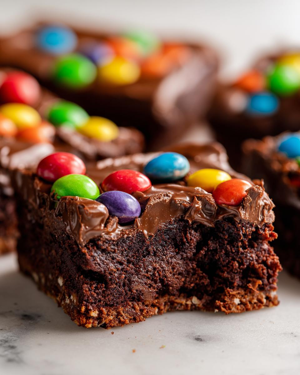 A close-up of a fudgy square of cosmic brownies topped with chocolate frosting and colorful candy-coated chocolates.