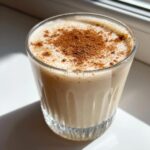 A glass of creamy, frothy coquito topped generously with ground cinnamon, sitting in bright sunlight.