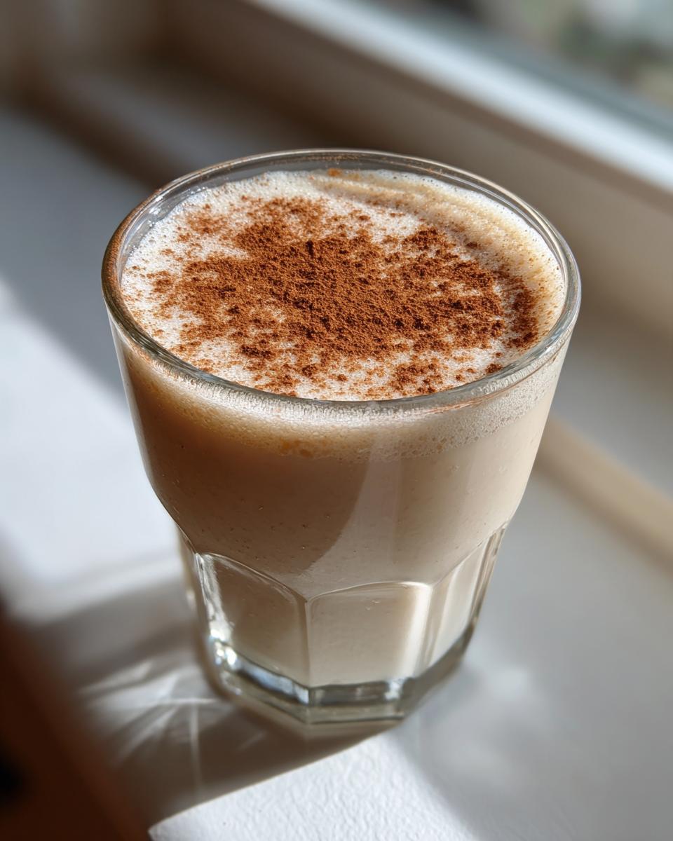 A glass of creamy, light brown coquito topped generously with ground cinnamon, sitting in natural sunlight.