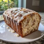 A freshly baked loaf of apple fritter bread drizzled heavily with white glaze, resting on a white plate.