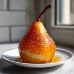 A single, glistening poached pear coated in a rich, amber glaze, sitting on a small white plate.
