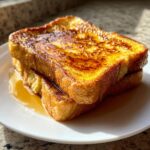 Close-up of two thick slices of golden brown french toast stacked and drizzled generously with maple syrup.