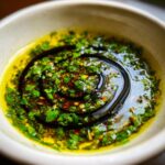 Close-up of a vibrant olive oil bread dip featuring bright green herbs and a swirl of dark balsamic vinegar.