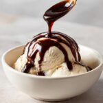 A spoonful of rich chocolate syrup being drizzled over scoops of vanilla ice cream in a white bowl.