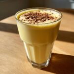 A tall glass filled with creamy yellow homemade eggnog, topped with dark chocolate shavings, sitting on a wooden surface.