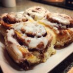 Four freshly baked cinnamon rolls dripping with white icing served on a white plate.
