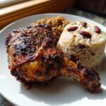 Close-up of a juicy, charred Jamaican jerk chicken leg quarter served with rice and peas.