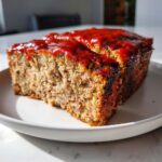 A thick slice of juicy meatloaf topped with a bright red glaze, served on a white plate.