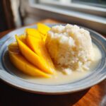 A serving of bright yellow sliced mango next to sweet sticky rice drenched in coconut sauce, creating delicious mango sticky rice.