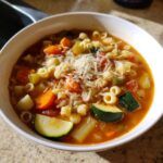 A close-up of a white bowl filled with rich minestrone soup, featuring vegetables, pasta, and grated Parmesan cheese.