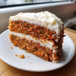 Close-up of a moist slice of carrot cake recipe featuring visible shredded carrots and thick cream cheese frosting layers.