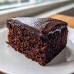 Close-up of a thick slice of moist chocolate cake topped with glossy dark chocolate frosting.