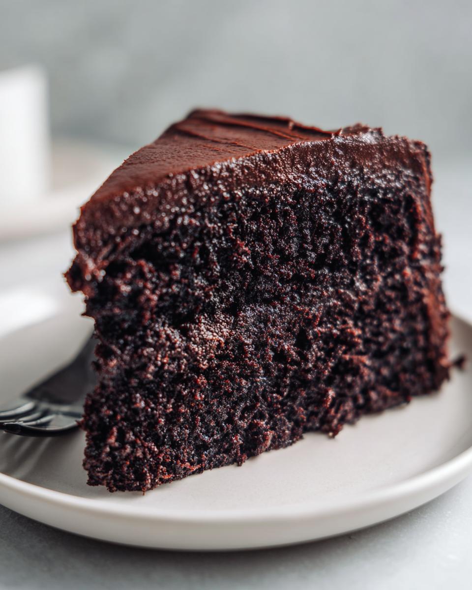 Close-up of a rich, dark slice of moist chocolate cake topped with thick chocolate frosting on a white plate.