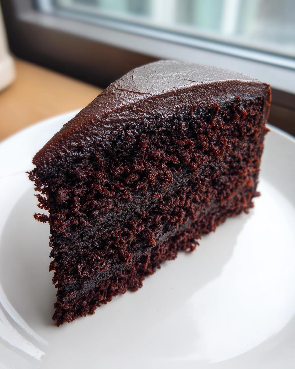 A close-up of a rich, dark slice of moist chocolate cake topped with glossy chocolate frosting.