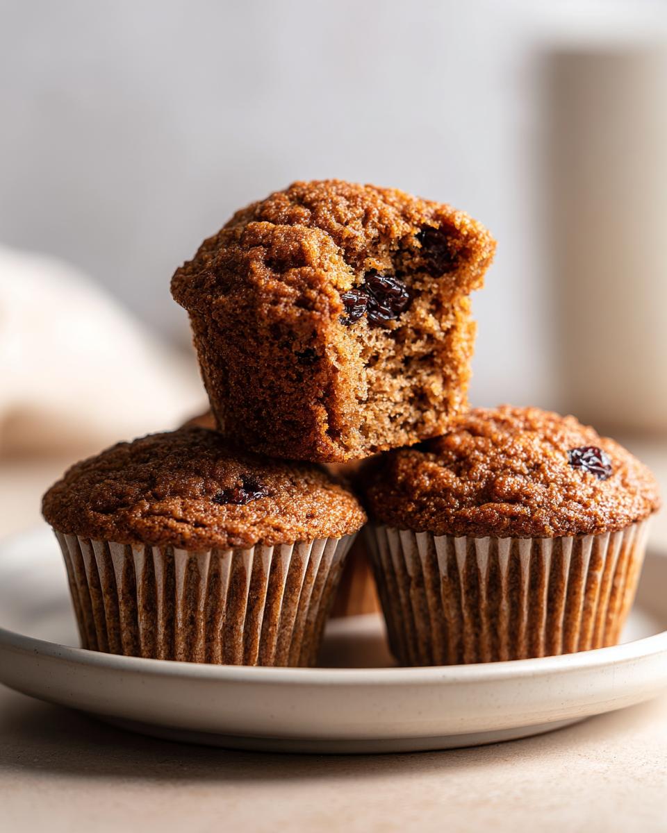 A stack of three moist honey bran muffins, one on top with a bite taken out showing the texture and raisins inside.
