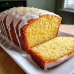 Close-up of a moist lemon loaf cake, partially sliced, drizzled with white glaze and topped with lemon zest.