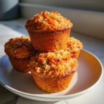 A stack of four golden brown morning glory muffins with visible shredded carrots and a crumbly topping, sitting on a white plate.