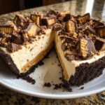 A rich slice cut from a no-bake butterfinger pie, showing layers of chocolate crust, peanut butter filling, and topped with chopped peanut butter cups.