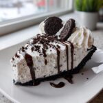 A decadent slice of cookies and cream ice cream pie with whipped cream, chocolate drizzle, and Oreo cookies.