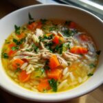 Close-up of a white bowl filled with hot penicillin soup featuring shredded chicken, bright orange carrots, orzo pasta, and fresh parsley.