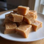 A stack of soft, light brown penuche fudge squares sprinkled with coarse sea salt on a white plate.