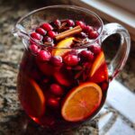 Close-up of a glass pitcher filled with vibrant red christmas sangria, garnished with fresh cranberries, orange slices, cinnamon sticks, and star anise.