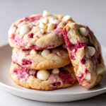 A stack of three soft raspberry cheesecake cookies featuring visible white chocolate chips and a fourth cookie broken open.
