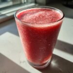 A tall glass filled with a bright red, icy watermelon smoothie, glistening with condensation in the sunlight.