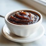 A close-up of thick, dark brown apple butter served in a small white bowl on a saucer.