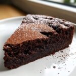 A decadent slice of flourless chocolate torte dusted with cocoa powder and powdered sugar on a white plate.