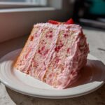 A tall slice of moist strawberry cake with pink frosting and visible strawberry pieces, sitting on a white plate.