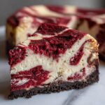 A close-up slice of rich red velvet cheesecake featuring vibrant red and cream swirls over a dark chocolate crust.