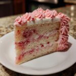 A close-up of a slice of light sponge rose cake with pink raspberry swirls and pink piped frosting, topped with gold flakes.