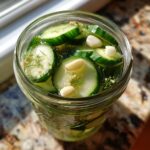 Overhead view of sliced cucumbers, dill, and garlic cloves being made into refrigerator pickles in a clear glass jar.
