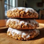 Three delicious iced oatmeal cookies stacked vertically on a wooden surface, covered in white vanilla glaze.