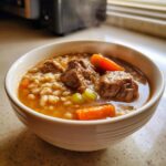 Close-up of a steaming white bowl filled with rich beef barley soup, featuring chunks of beef, barley, and carrots.