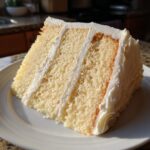 Close-up of a tall slice of moist vanilla cake recipe layered with thick white buttercream frosting.