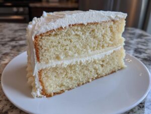 A perfect slice of moist vanilla cake recipe, showing two layers separated by white frosting, served on a white plate.