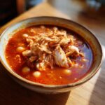 A close-up bowl of rich red chicken pozole, topped with shredded chicken and hominy kernels.